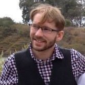 A portrait of a smiling, bearded man in a vest and checkered shirt.